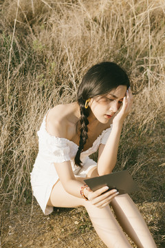白衣黑辫女子坐在干燥的草原上，在阳光明媚的日子里举着方镜。