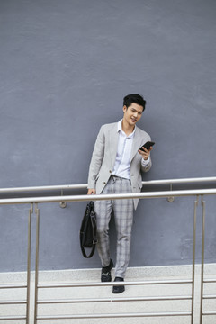 聪明的泰国亚裔商人穿着正式的灰色西装，手持智能手机，背景是灰色的。