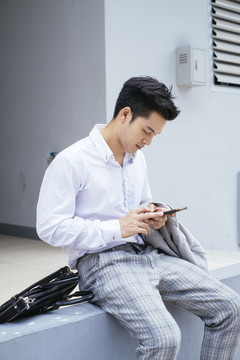 聪明的亚泰商人穿着白领衬衫坐在户外使用智能手机。