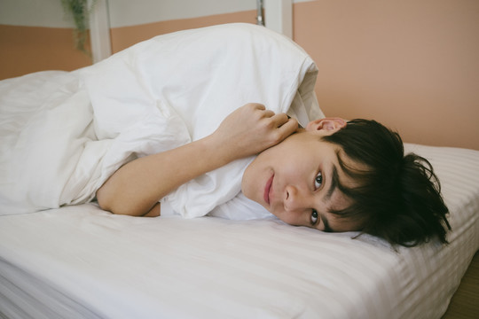 性格内向的家伙在休息日躺在床上，拿着白毛毯，看着镜头。
