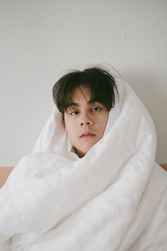 泰国男孩坐在床上，用白色毯子裹着身体。他用诱人的目光直视前方。