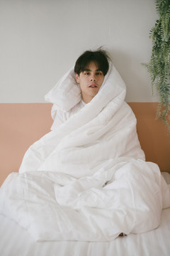 黑发男坐在墙边的床上，用白毯子裹着自己，没有表情。