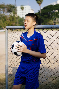 一个穿着蓝色足球服的男孩站在篱笆边拿着球。
