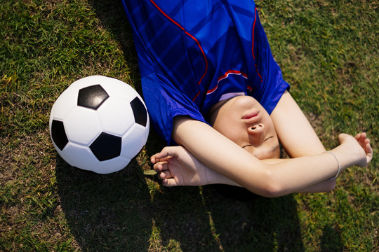 一个穿足球服的男孩躺在球场上，边睡边拿球。