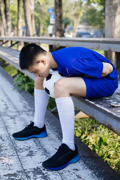 坐在看台上的小男孩两腿间夹着一个球，弯下身子。