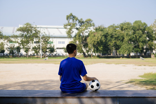 一个男孩坐在木凳上，手里拿着足球看着球场。