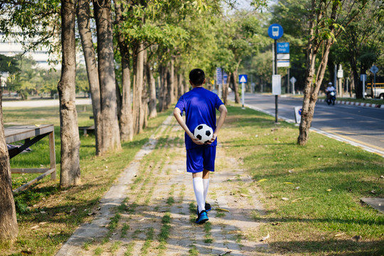一个男孩背着足球走在公园的人行道上。