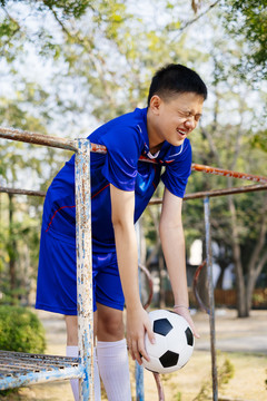 穿着蓝色套装的快乐男孩站在操场上的攀爬架上，踢着足球。