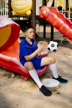 一个小足球运动员坐在操场上玩他手里的球。