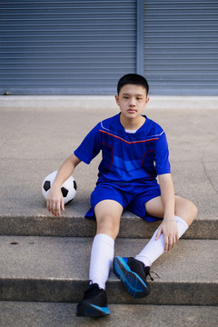 一个穿着蓝色足球服的男孩坐在鹅卵石楼梯上踢足球。