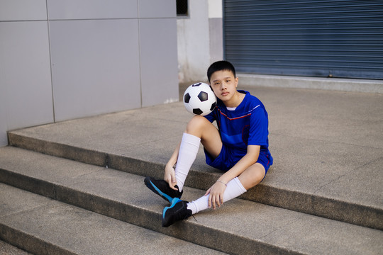 一个穿着蓝色足球服的男孩坐在鹅卵石楼梯上踢足球。