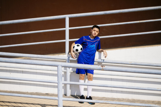 一个男孩靠在斜坡小路的扶手上，胳膊上抱着足球。