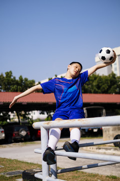 一个疯狂的男孩以危险的姿势坐在扶手上踢足球。以蓝天为背景。