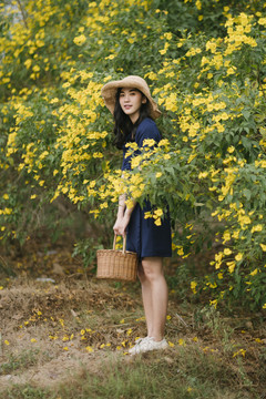 身穿深蓝色连衣裙的黑色长发女孩站在黄色的花丛里提着篮子。