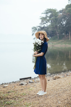全长美丽的泰国女孩站在湖边，抱着白色的花朵看着镜头。
