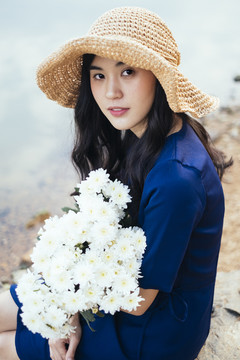 一个穿着深蓝色裙子的可爱女孩坐在湖边，怀里抱着一朵白花。