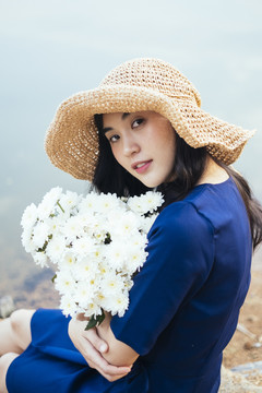 穿着深蓝色连衣裙的可爱女孩坐在湖边，抱着一朵白花。