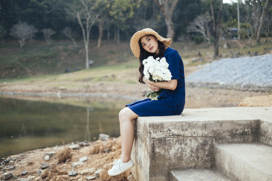一个穿着深蓝色衣服的漂亮女孩，抱着一朵白花坐在湖边的楼梯上。