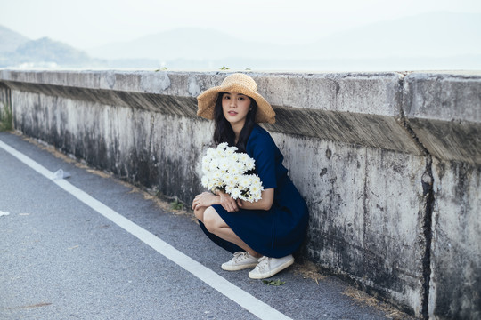 穿蓝衣服的女孩坐在路障旁，手里拿着白花，看着摄像机。