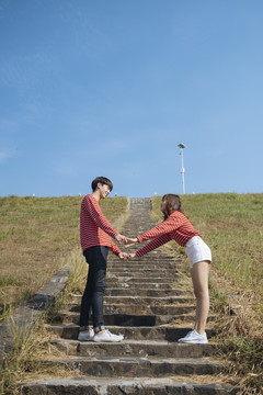 一对年轻的情侣站在石阶上，在蓝天下互相牵手。