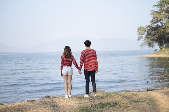 一对年轻夫妇站在湖边，握着对方的手，望着远方。