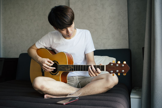 一位年轻的泰国吉他手一边用耳机听智能手机里的歌曲，一边演奏原声吉他。