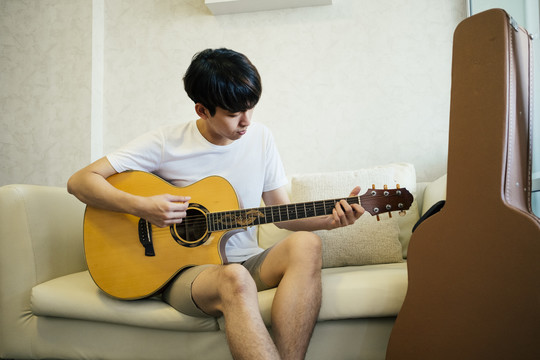 年轻的泰国吉他手在家客厅的沙发上弹原声吉他。