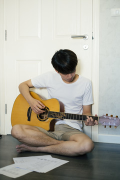 一个年轻的泰国吉他手坐在地板上在家里练习原声吉他。