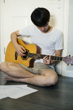 一个年轻的泰国吉他手坐在地板上在家里练习原声吉他。