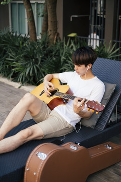 年轻的泰国吉他手用耳机听音乐，在沙滩椅上弹原声吉他。