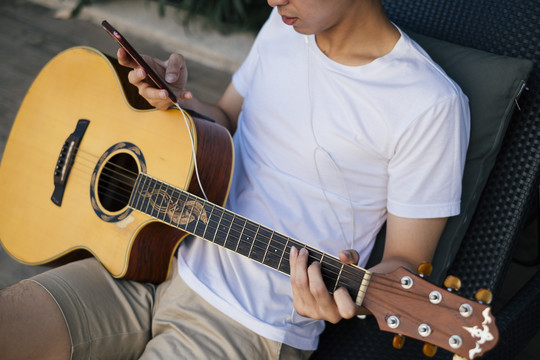 年轻的泰国吉他手用耳机听音乐用智能手机练习吉他。