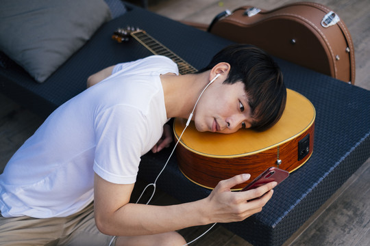 年轻的泰国吉他手躺在吉他上用耳机听智能手机里的音乐。