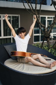 一个年轻的泰国吉他手在沙发上醒来，腿上放着原声吉他。和吉他睡觉。带着吉他醒来。