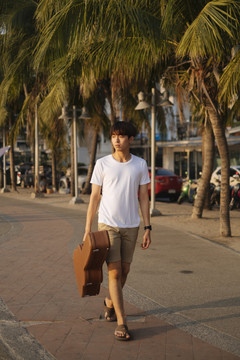 一个年轻的泰国吉他手手手拿着吉他盒在日落时分的海滩上散步。