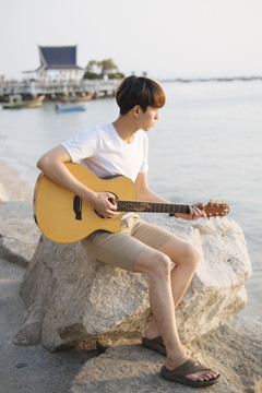 年轻的泰国吉他手男子坐在岩石上弹吉他在海滩上日落。