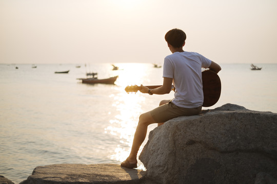 后视图-剪影年轻的泰国吉他手男子在海滩上弹原声吉他在日落时间。