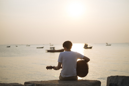 后视图-剪影年轻的泰国吉他手男子在海滩上弹原声吉他在日落时间。