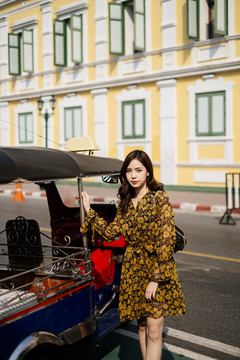 身着黄色连衣裙的可爱卷发女孩站在粉彩大楼前的马路上，抢着tuktuk汽车。