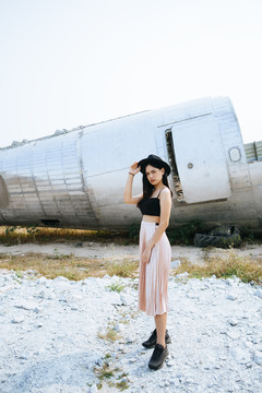 黑色长发女孩穿着黑色意大利面皮带和粉色裙子站在废弃区与飞机残骸。