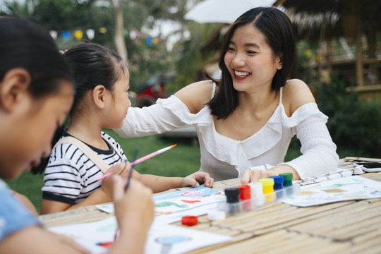 快乐的亚洲大姐抚摸着小妹妹的头表示恭维。两姐妹在户外公园的纸上画水彩。