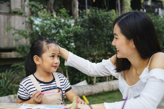 快乐的亚洲大姐抚摸着小妹妹的头表示恭维。两姐妹在户外公园的纸上画水彩。