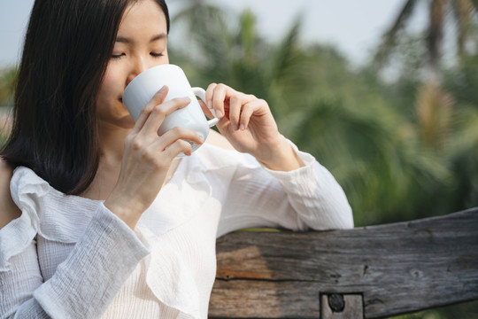 一位泰国亚裔妇女坐在乡村地区的木凳上喝咖啡。在大自然中旅行。在一杯咖啡中加香。喝白咖啡的漂亮女人。