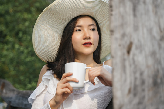 泰国亚裔女子戴着帽子和白衬衫坐在木制长凳上喝咖啡在农村地区。在大自然中旅行。在一杯咖啡中加香。喝白咖
