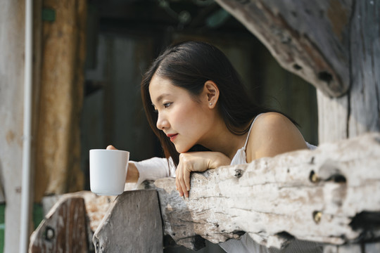 在乡村旅馆的木制阳台上，一名亚泰妇女拿着一杯咖啡向外眺望。