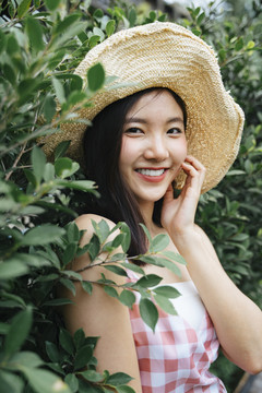 在一片绿叶植物旁，一位身穿粉色连衣裙、戴着帽子、面带微笑的快乐亚洲女孩的特写镜头。