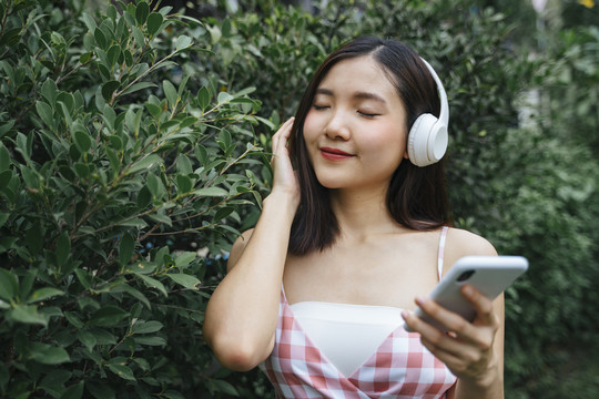 戴着耳机的美丽年轻女子站在树上，闭着眼睛微笑着听音乐。