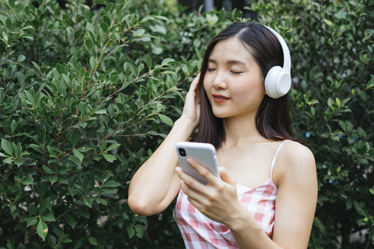 戴着耳机的美丽年轻女子在绿树旁的花园里闭着眼睛微笑着听音乐。