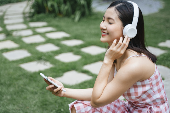 戴着耳机的美丽年轻女子坐在绿树旁的花园里，微笑着听着音乐。