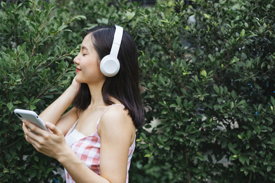 侧视图-戴着耳机的美丽年轻女子在绿树植物附近的花园里，闭着眼睛微笑着听音乐。