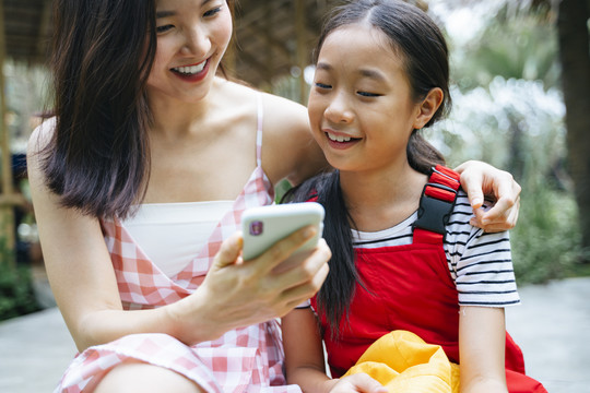 快乐的两个亚洲姐妹坐在公园里，一起玩智能手机，一起笑。